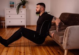 A bearded man in black fitness attire exercising at home, showcasing arm tattoos and a healthy lifestyle.