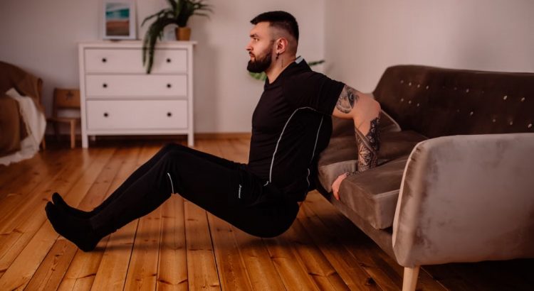 A bearded man in black fitness attire exercising at home, showcasing arm tattoos and a healthy lifestyle.