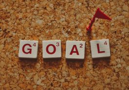 Scrabble tiles forming the word 'goal' with a small red flag on a cork board background.